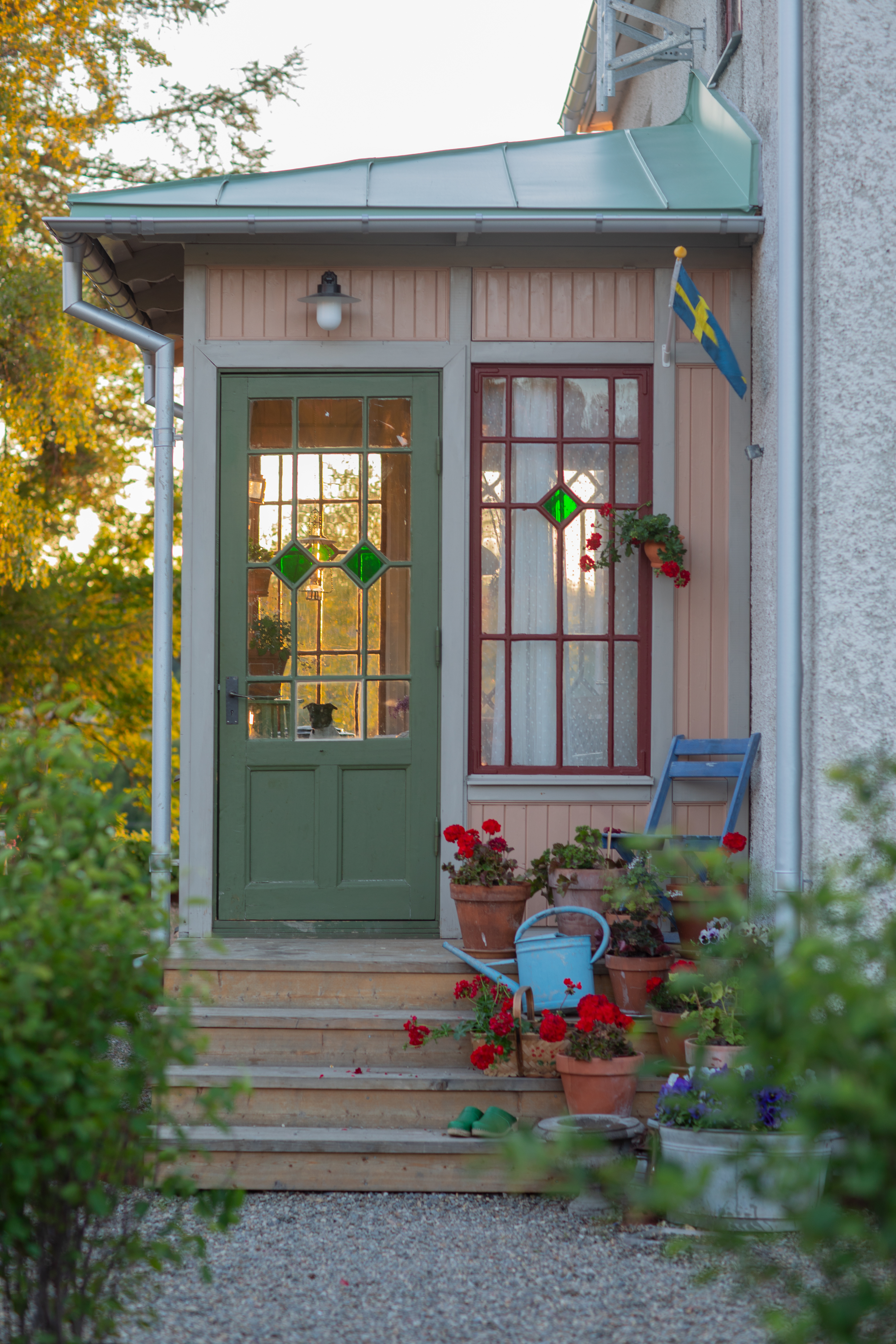Entré till ett hus med en grön dörr och färgade glasrutor. På verandan finns blomkrukor med röda och gröna växter, en blå vattenkanna, och en svensk flagga hänger intill.