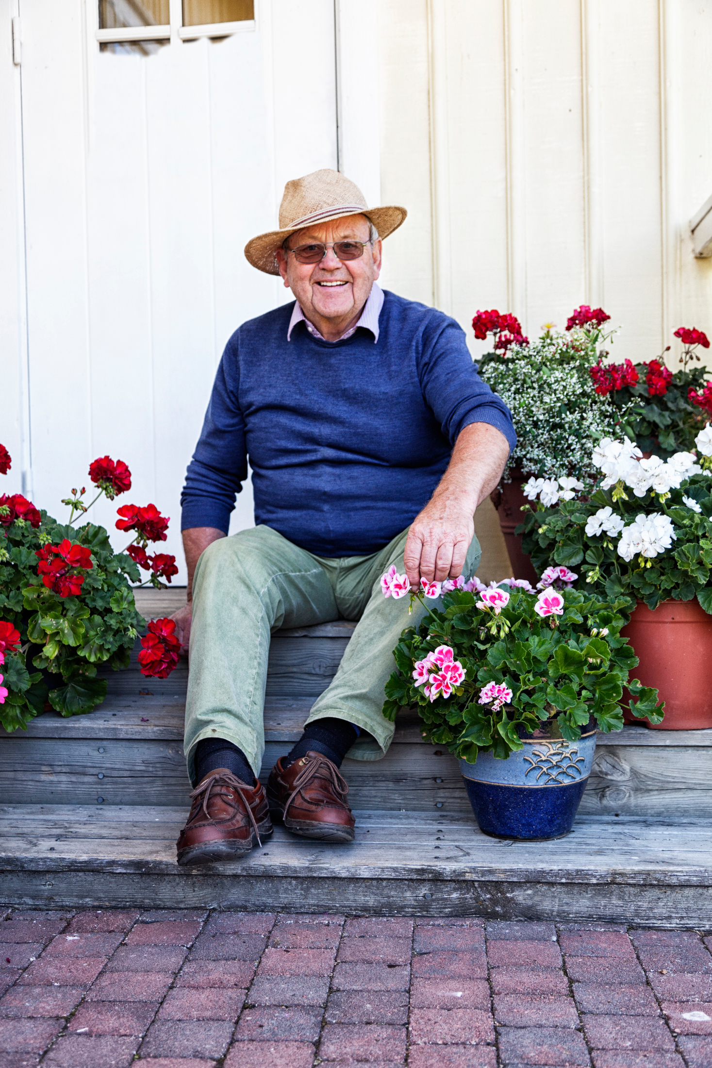 En äldre man i blå tröja och halmhatt sitter leende på en trappa, omgiven av blommande pelargoner i krukor.